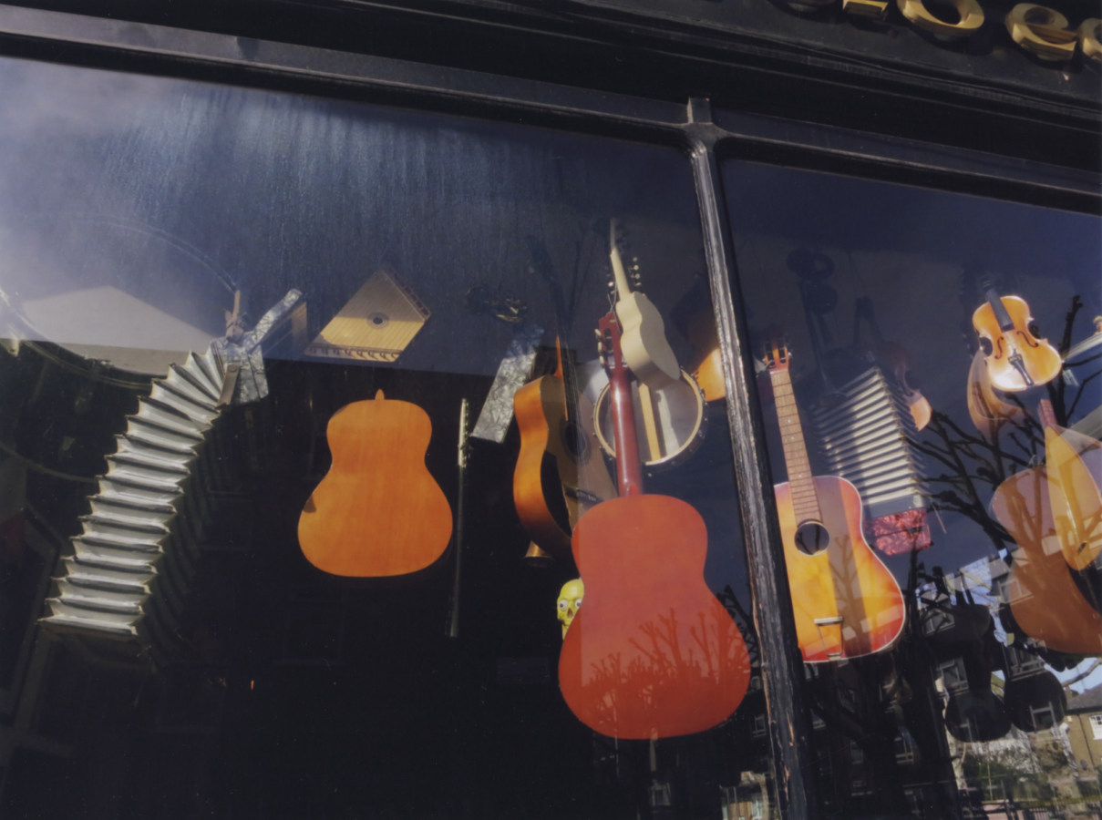 A color photograph of many musical instruments (mostly acoustic guitars), hanging in a storefront window