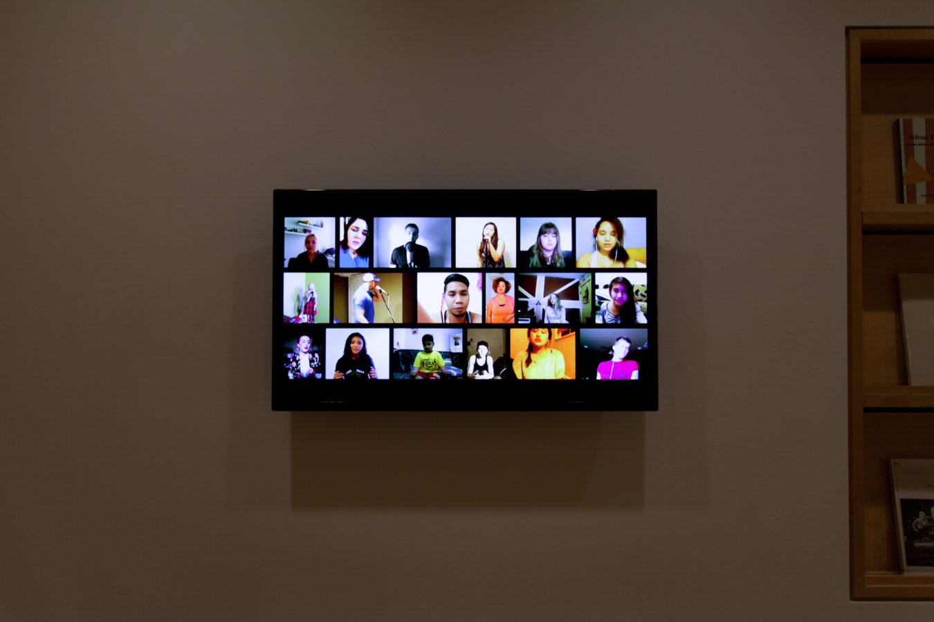 Color image of video work on TV screen depicting multiple people singing on white gallery wall