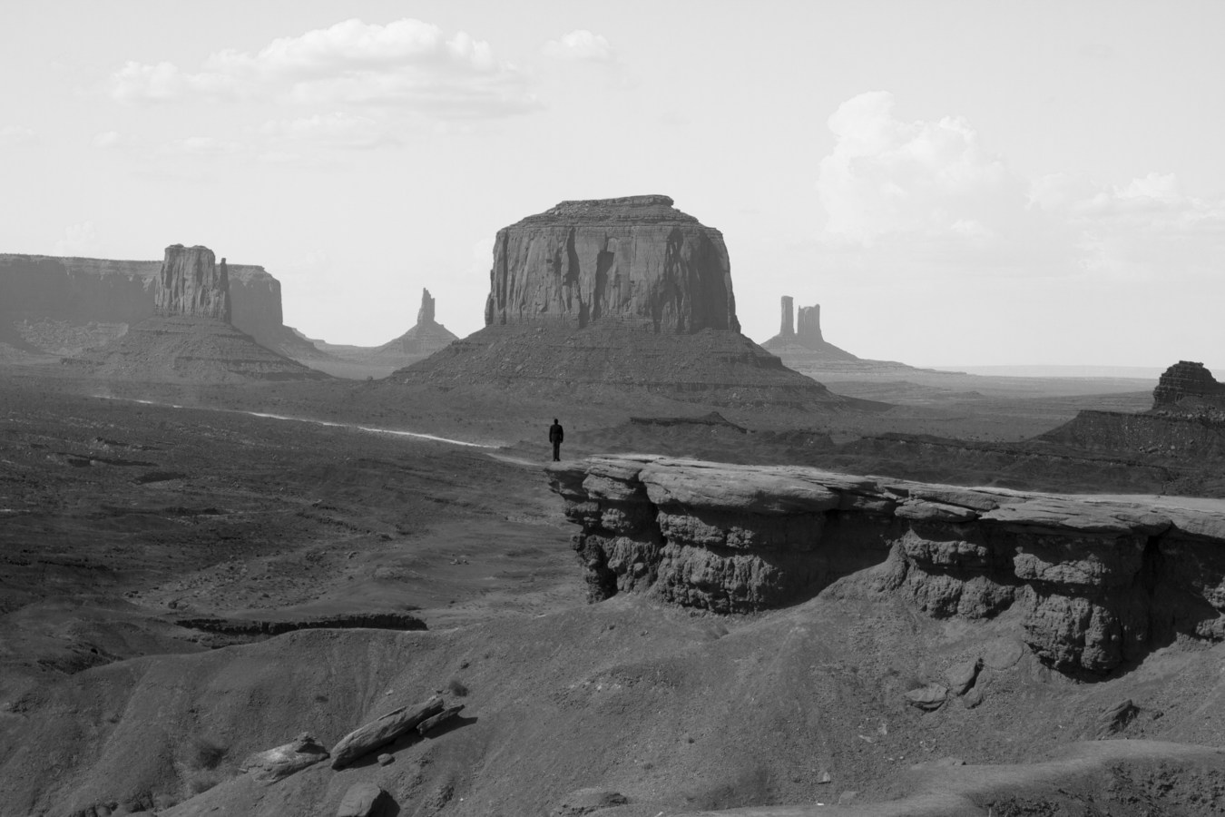 Black and white photograph of lone silhouetted figure in vast landscape of John Ford Point, in Monument Valley.