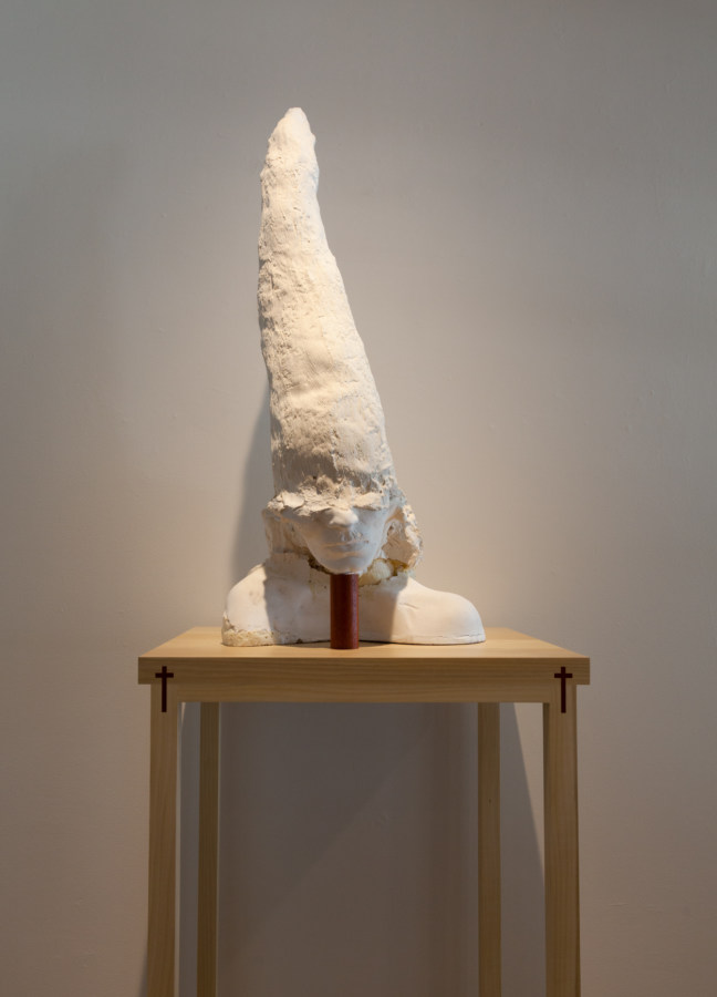 Color image of white sculpture depicting a head adorned with large hood on wooden stand with crucifixes