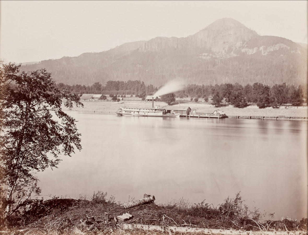 Ninteteenth century photograph of a steam ship at a pier on a river with a mountain in the background