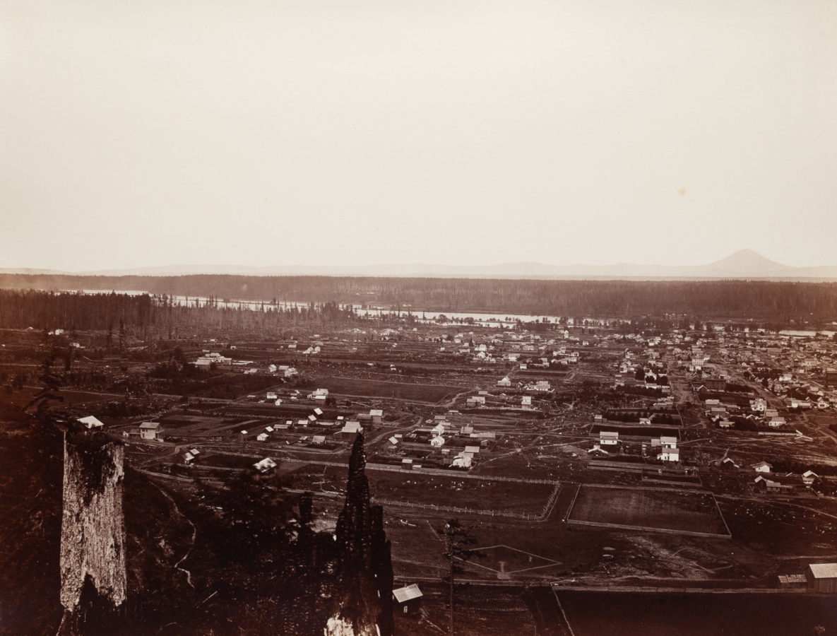 Ninteteenth century photograph of faraway buildings and houses, with a river behind them, from an elevated vantage point