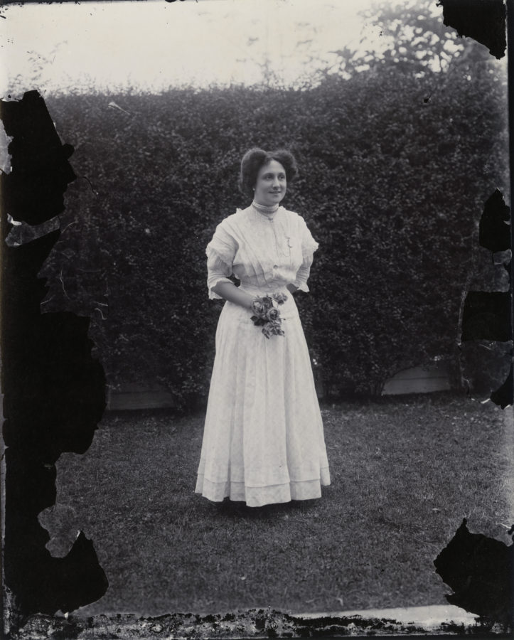 Black and white photograph of a woman in a white dress standing outside in front of a hedge.