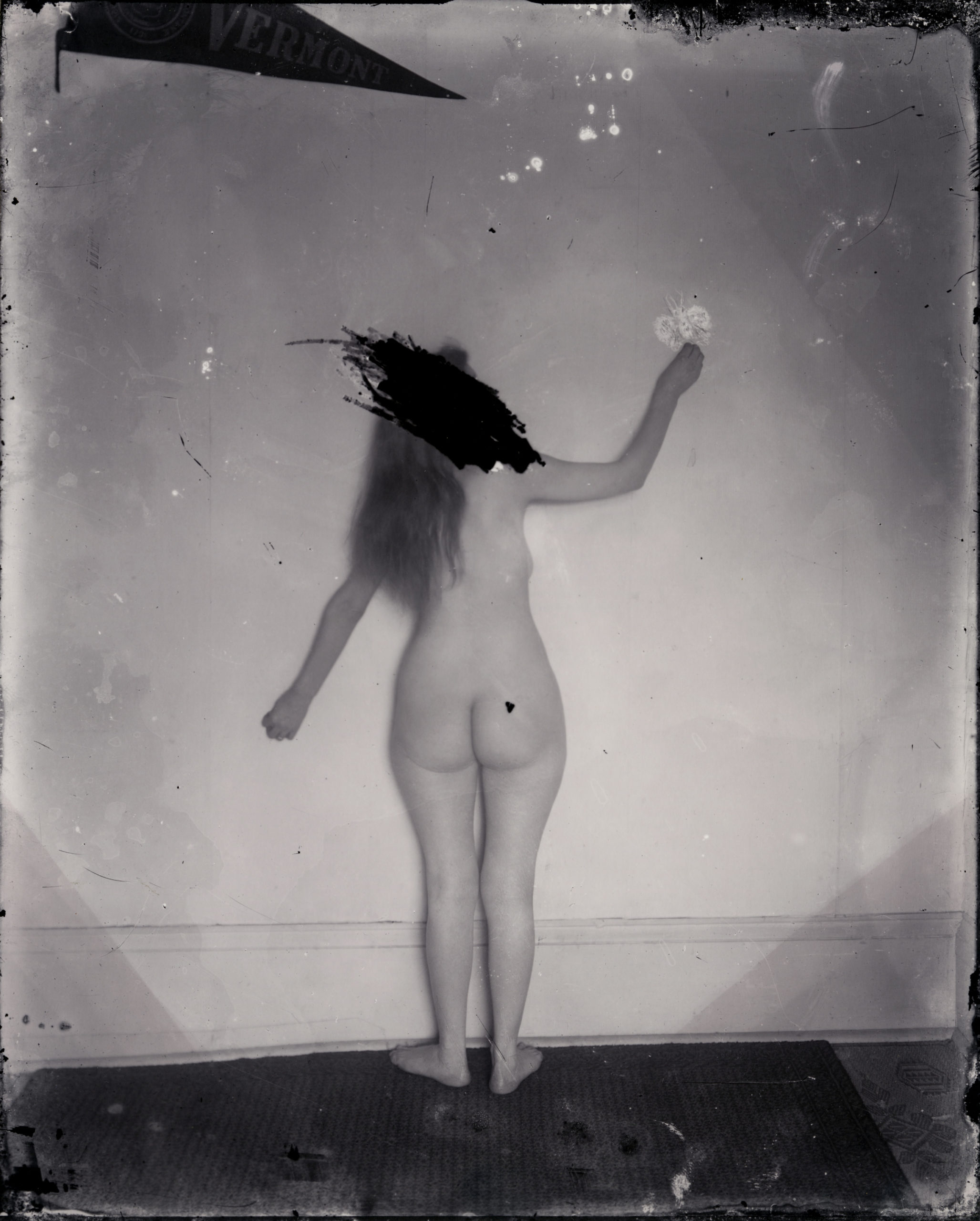 Black and white photograph of a nude woman facing a wall, with her face scratched out