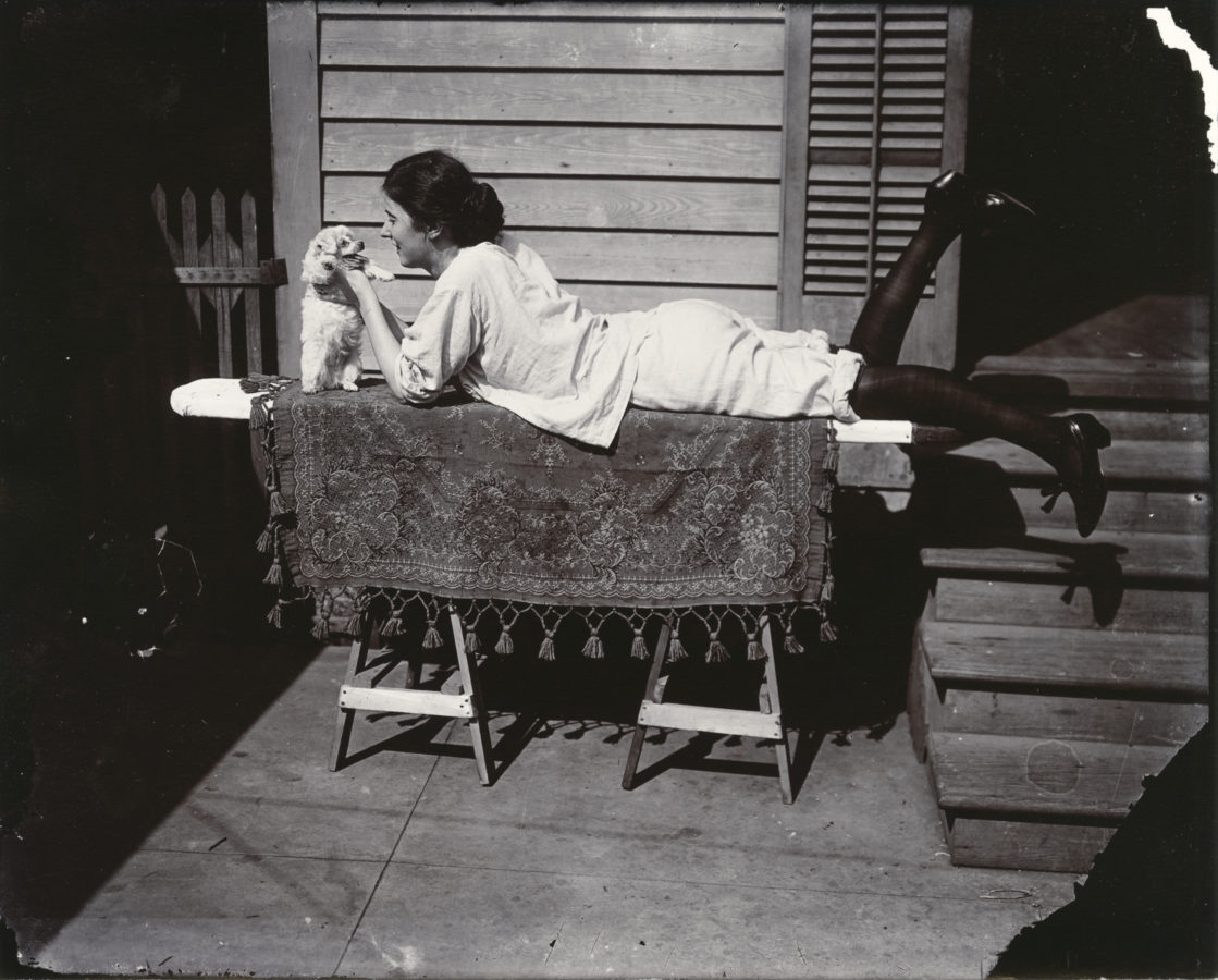 Black and white photograph of a woman laying on an ironing board, petting a small dog