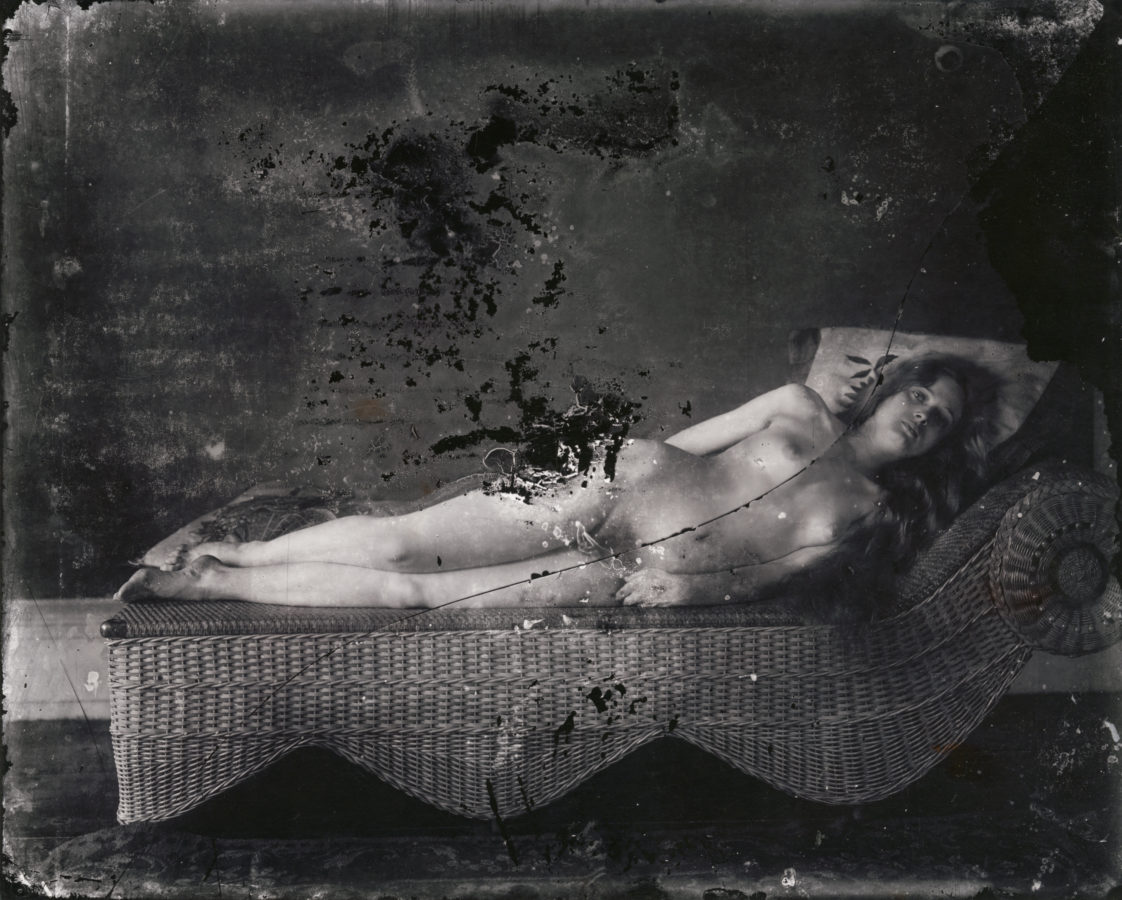 Black and white photograph of a nude woman reclining on a wicker lounge chair