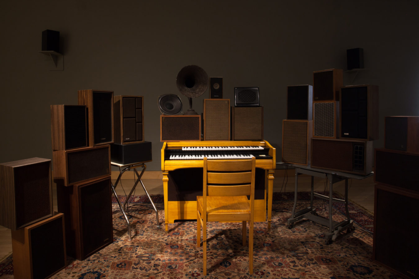 Photograph of an installation of a chair in front of a keyboard, surrounded by multiple stacked speakers