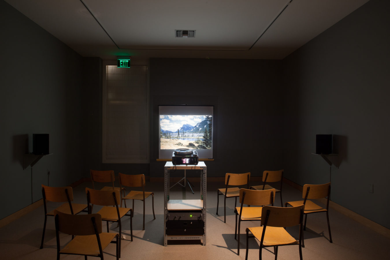 Photograph of an installation of a slide projector, flanked by chairs, projecting a color landscape onto a screen