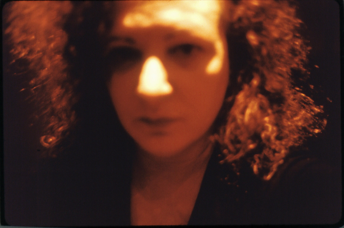 Color photograph of an out-of-focus woman's face framed by frizzy hair under orange light