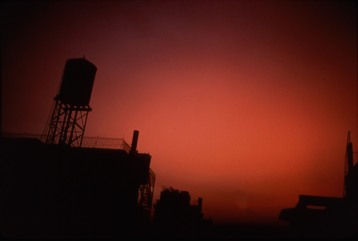 Color photograph of silhouetted industrial buildings against a red sky