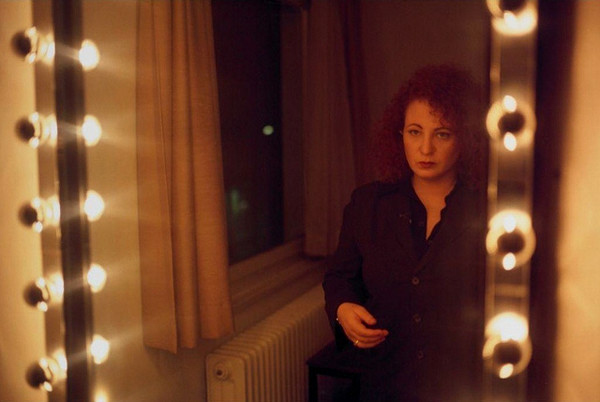 Color photograph of a mirror with circular lights on the sides, reflecting a woman standing, gazing into the mirror