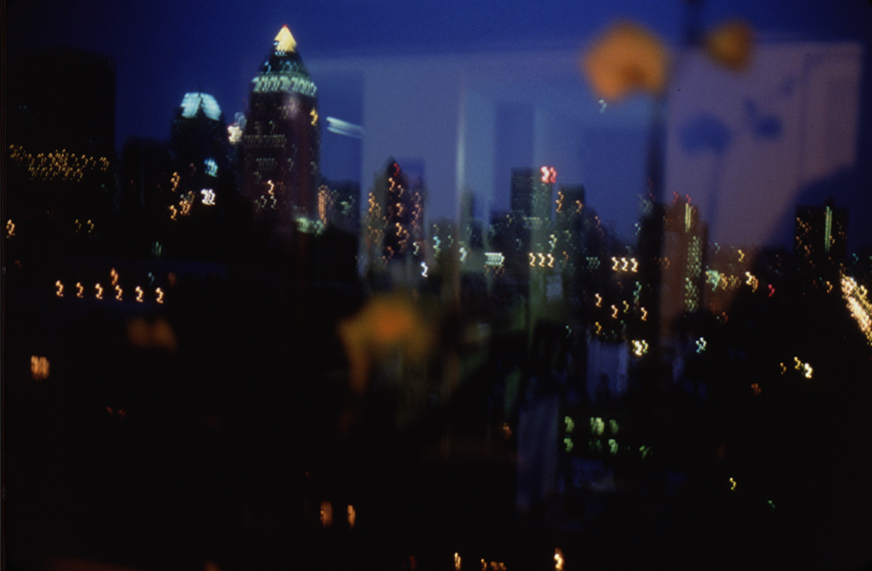 Color photograph from behind a window of an out-of-focus city skyline at night with faint reflections from the room superimposed