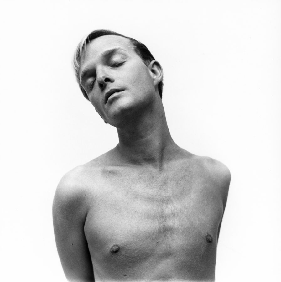 Black and white photograph of a young man, shirtless, with his eyes closed, against a white background.