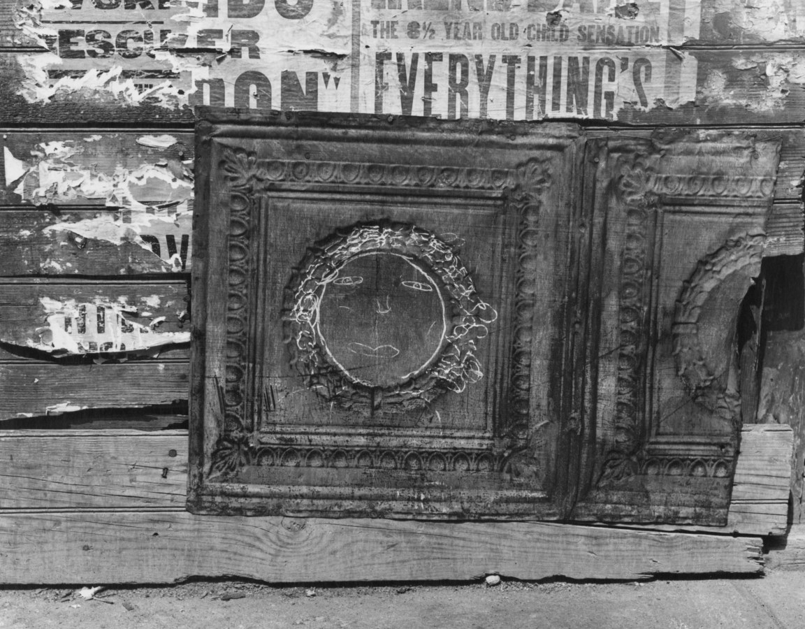 Black-and-white photograph of a wall with torn advertisements, and a moulded tin panel with a chalk drawing of a face.