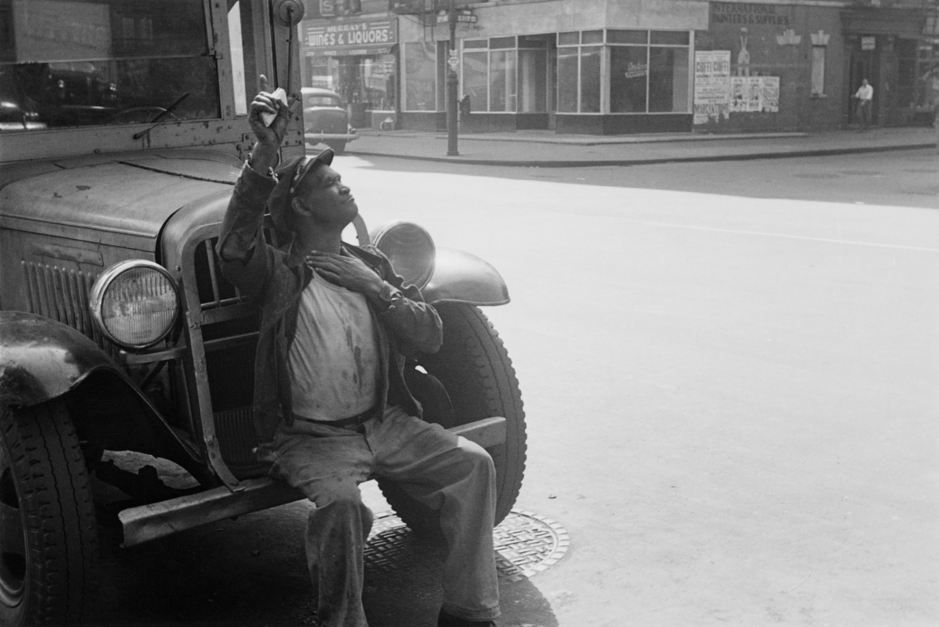 Black-and-white photograph of an African American man seated on the front bumper of a parked car.