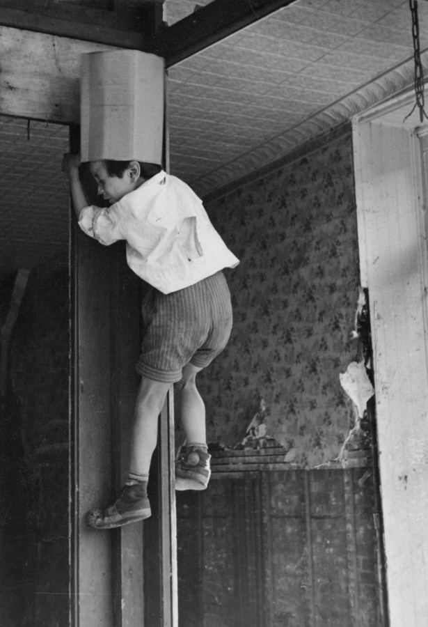 Black-and-white photograph of a young boy climbing a wall in a disheveled hallway.