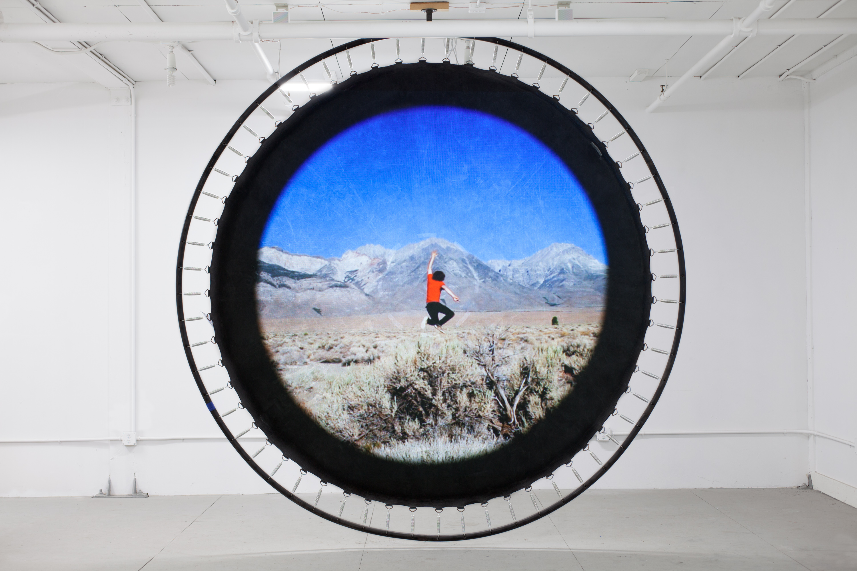 Trampoline hanging from a gallery ceiling with the image of a person jumping in a landscape projected onto it