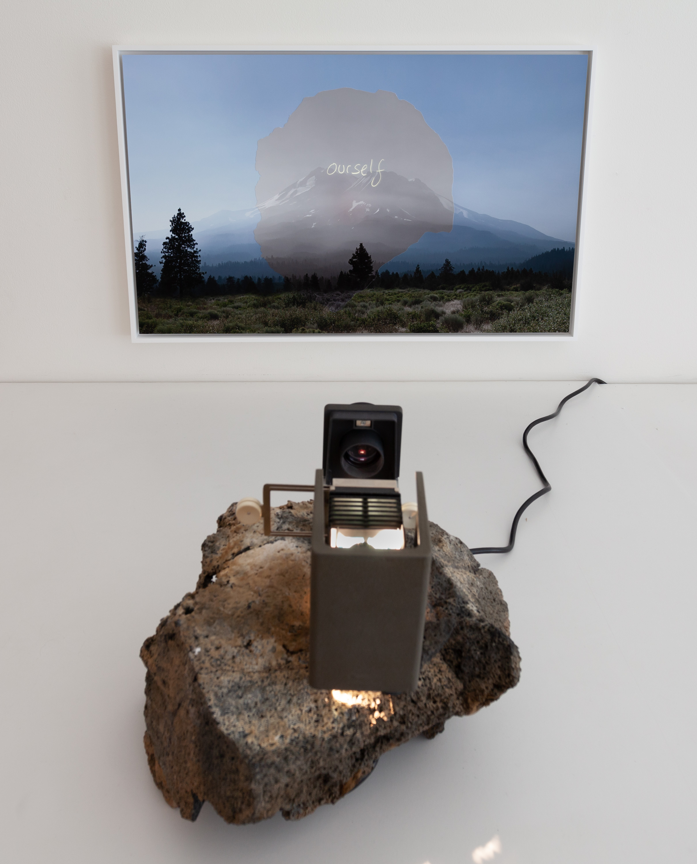 Color photograph of a mountain with overlaid text reading "ourself" being projected onto the image by a projector resting on a rock