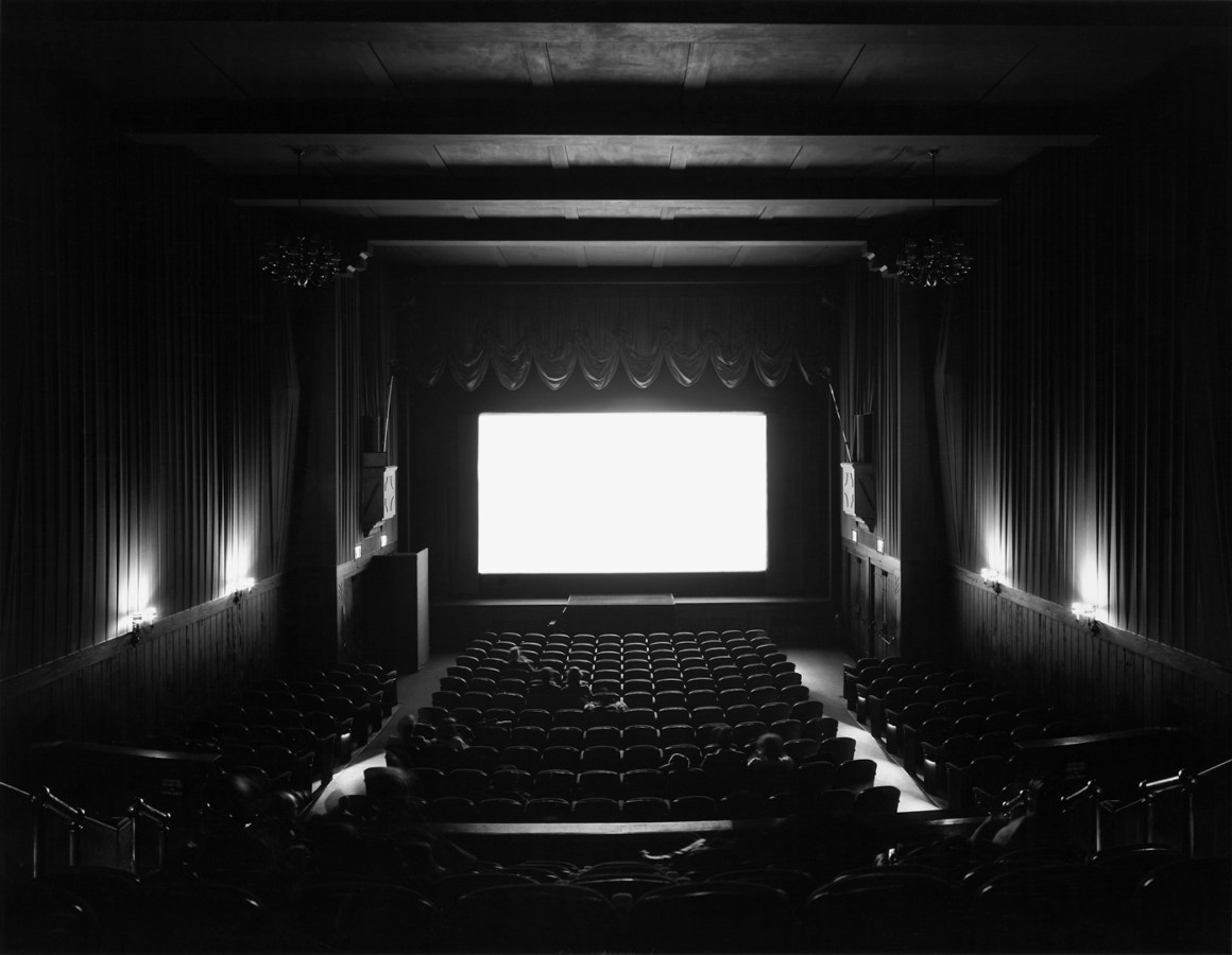 A black and white photograph of a movie theater with a bright white screen, and audience watching the film
