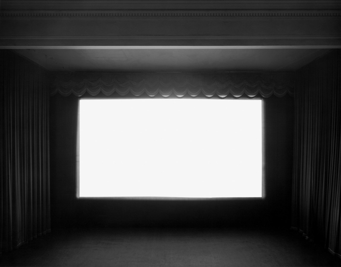 A black and white photograph of a movie theater with a bright white screen, with curtains on either side and at the top