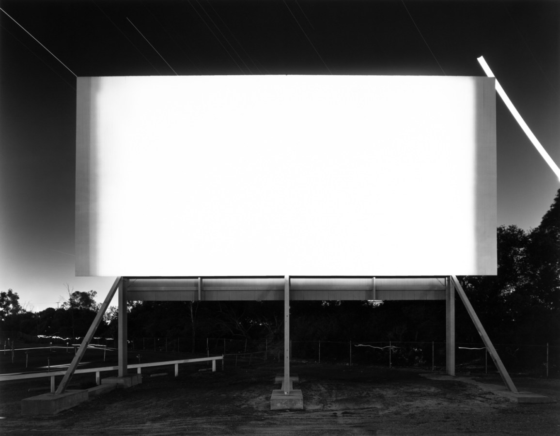 A black and white photograph of a bright white movie screen at a drive-in theater at night, with bright star trails behind it