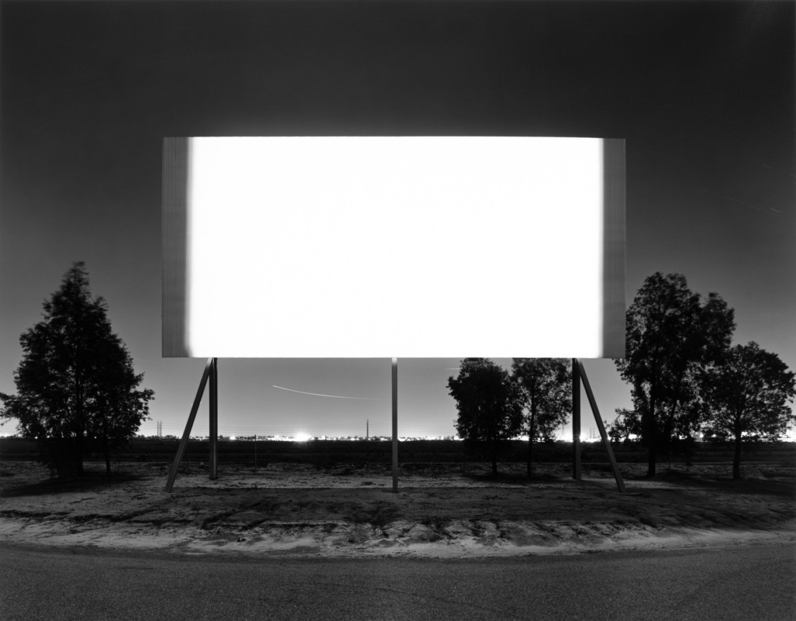 A black and white photograph of a bright white screen at a drive-in, with trees behind it and a city in the distance