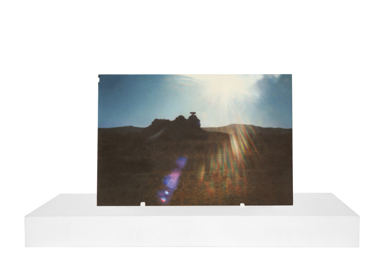 A painting of a vernacular photograph, on a white stand, of a rock formation out in the desert. The sun has caused a rainbow light flare in the center of the image.