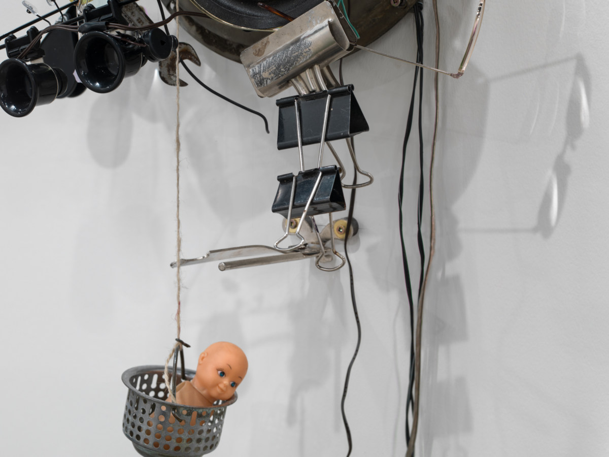Close-up installation view of a mixed-media sculpture, showing binder clips and a small toy.