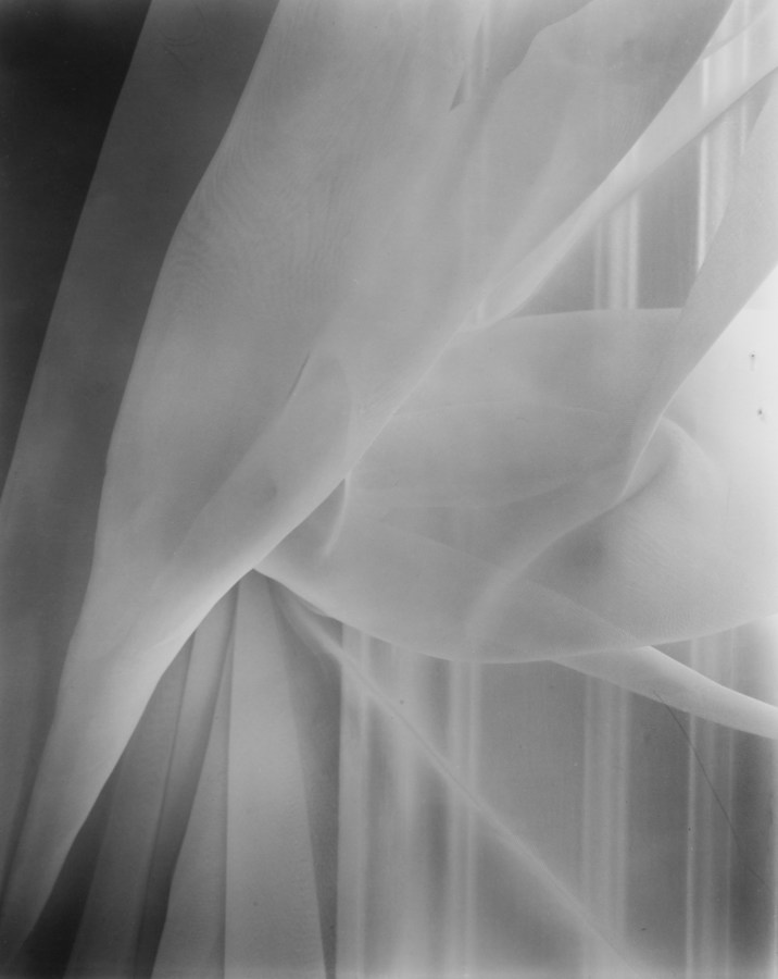 Black-and-white photograph of a window curtain pulled back onto a hook in the window frame