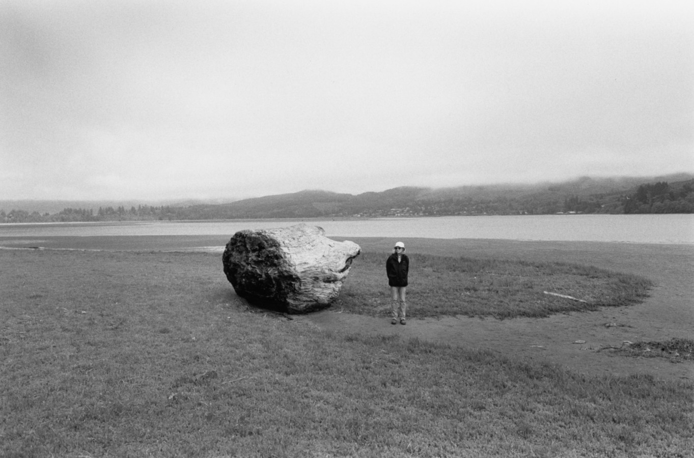 Black-and-white photograph of a woman standing next to a large driftwood stump.