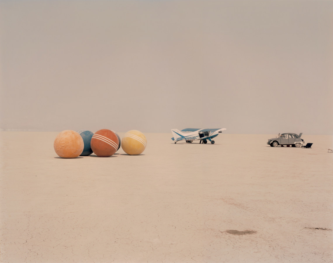 Color photograph of a desert field with a car and small aircraft, and four oversized croquet balls.
