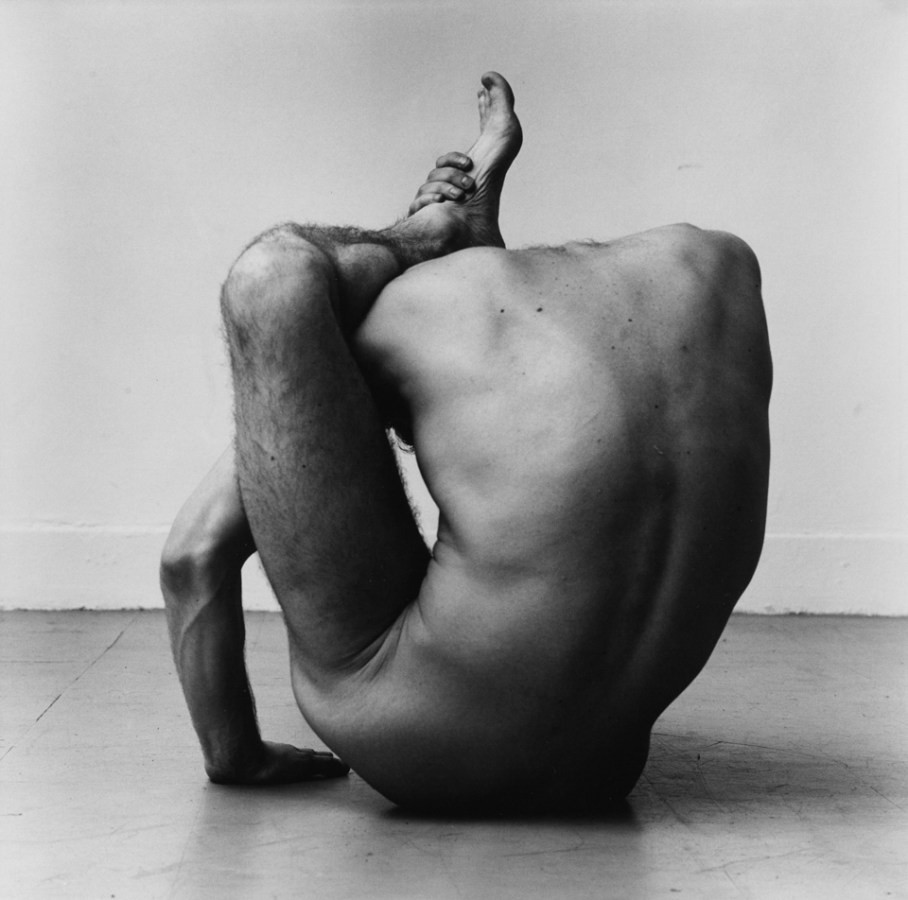 Black-and-white photograph of a nude man with his ankle hooked over his neck, seen from the back.