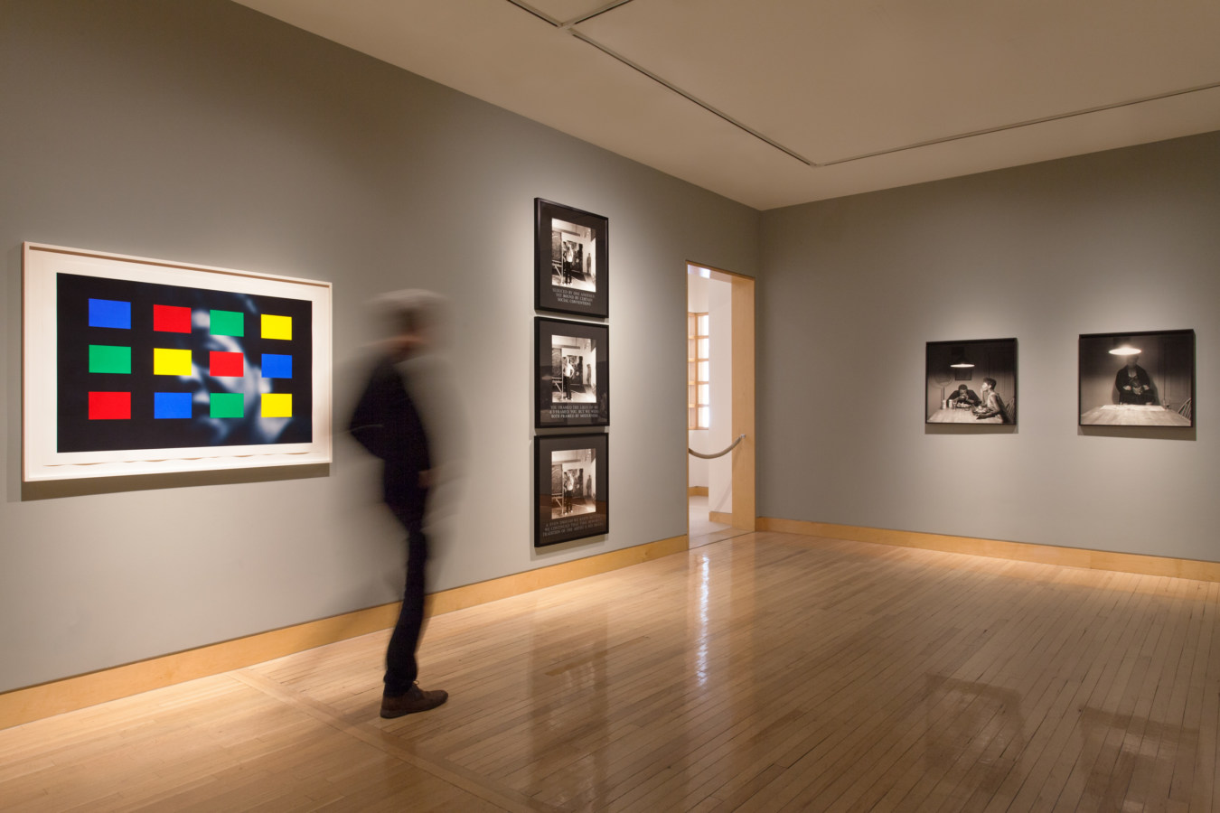 Color image of framed prints on grey gallery walls with person walking