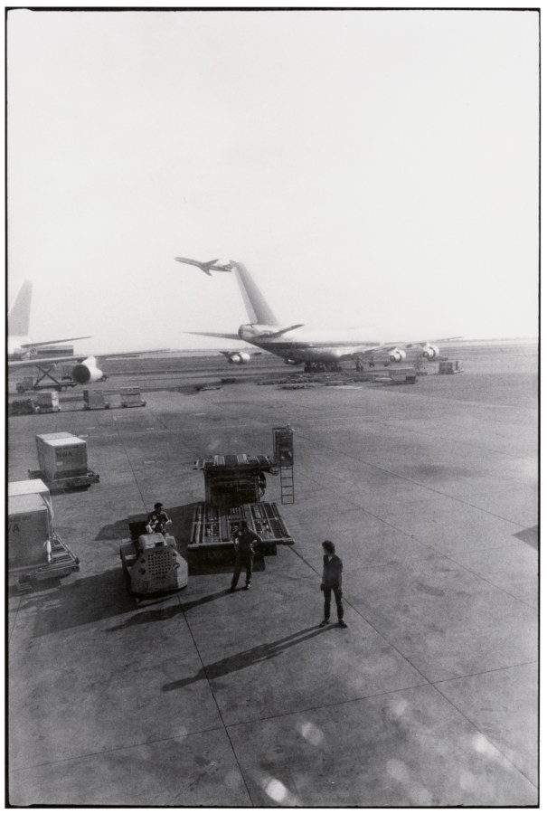 A black and white vertical photograph, taken from an airplane window while at the gate. Three men stand outside on the tarmac, with a plane in the background.