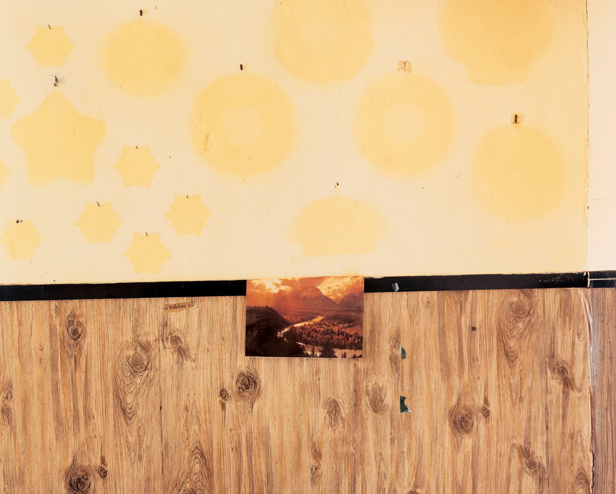 Color photograph of a post-card sized picture of a mountain river tacked to a wall of wood paneling and yellow patterned wallpaper