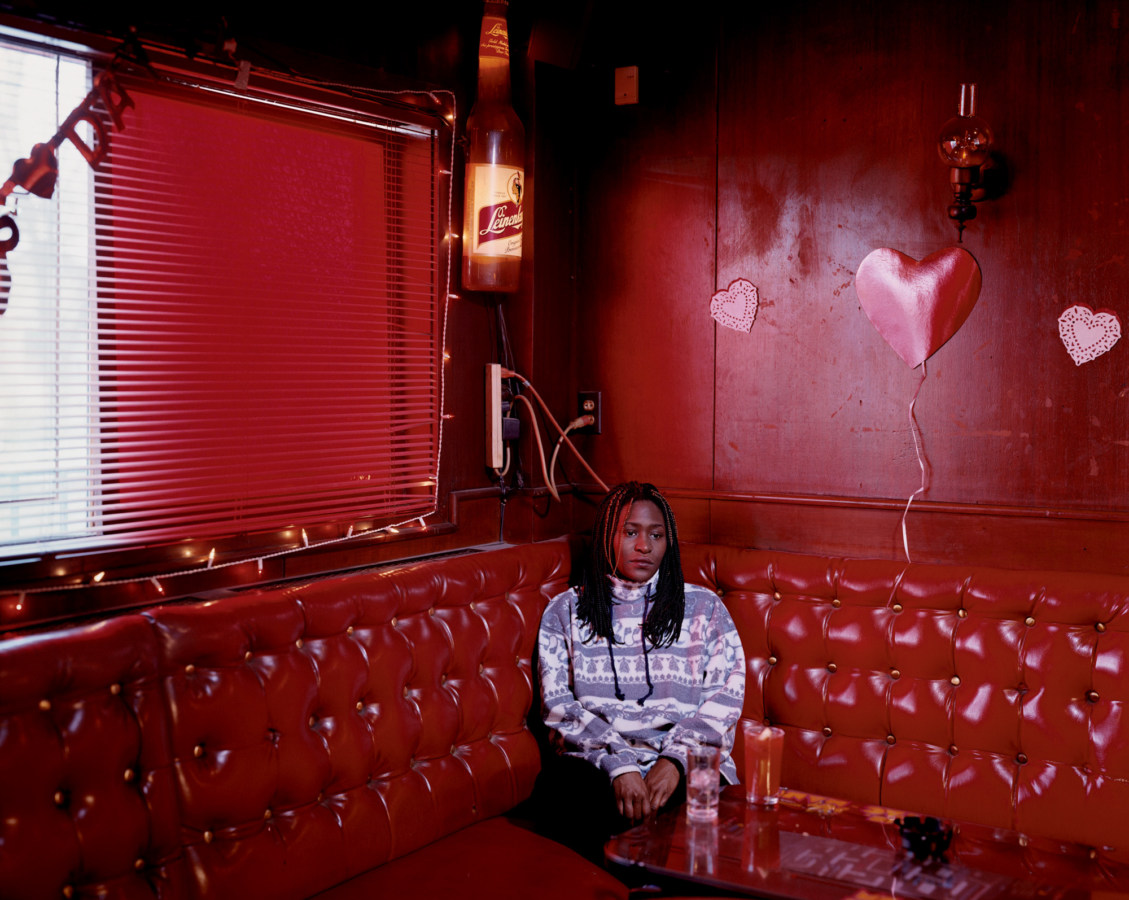 Color photograph of a young woman sitting in a red corner booth in an all-red room with paper hearts on the wall