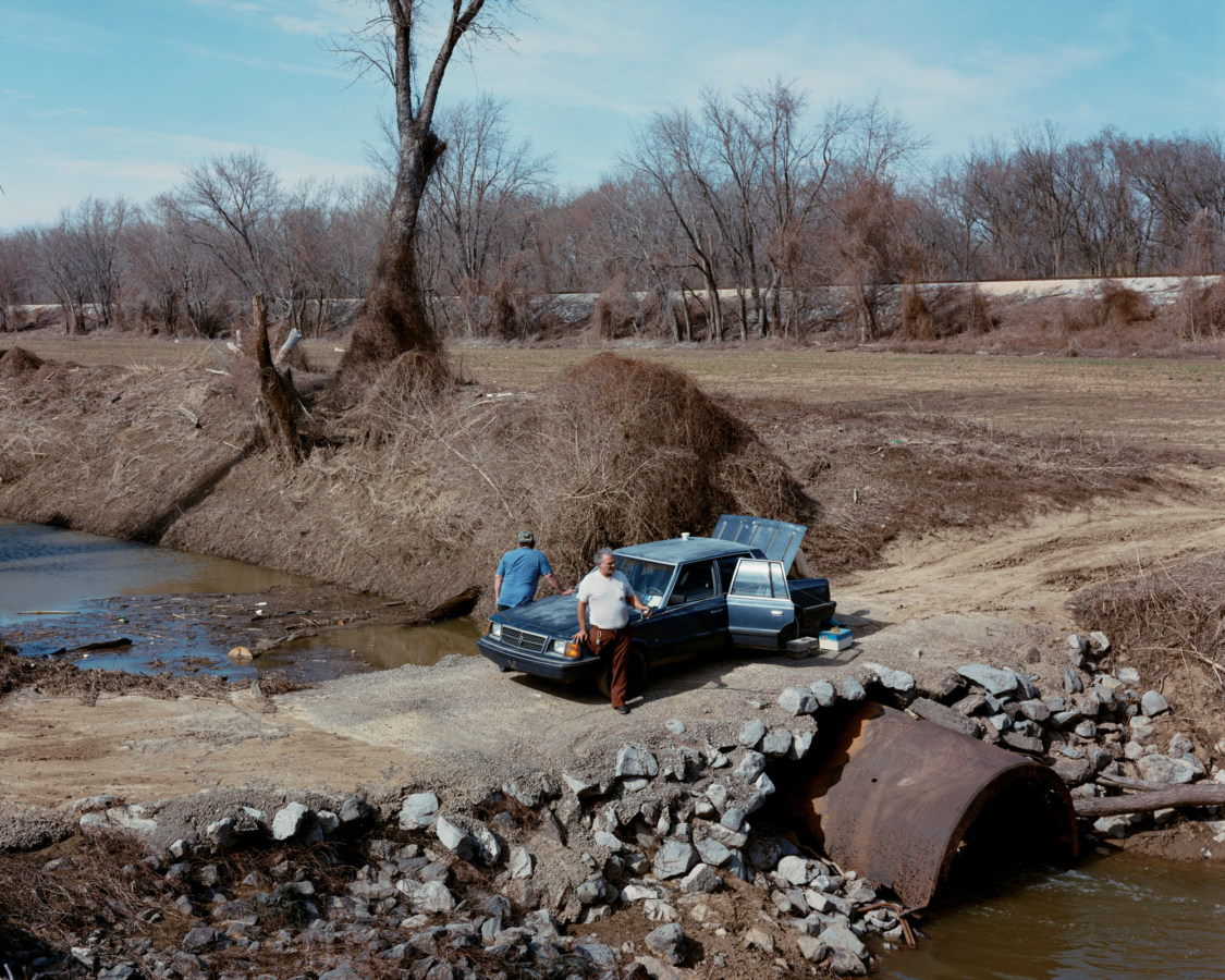 Color photograph of two men leaning on the hood of a car parked on a crossing over a wide ditch