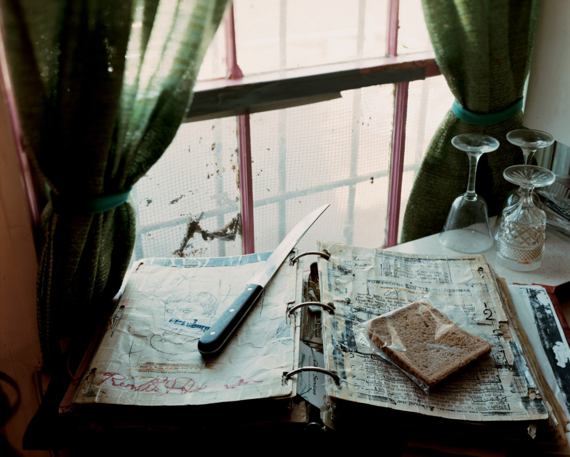 A color photograph of a book in front of a window with green curtains. On top of the book is a long knife and a piece of brown bread in a plastic bag.