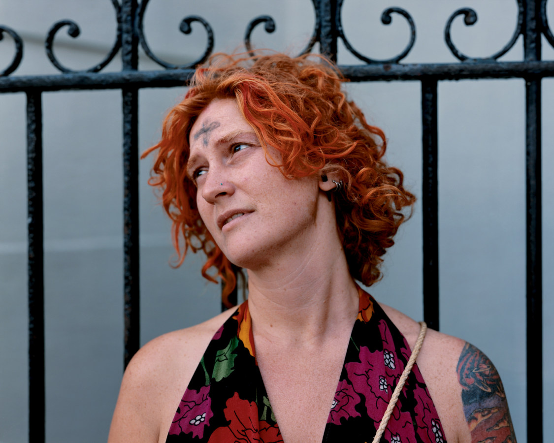 A color photograph of a white, red-haired woman in front of a fence. She has a black ash cross on her forehead.