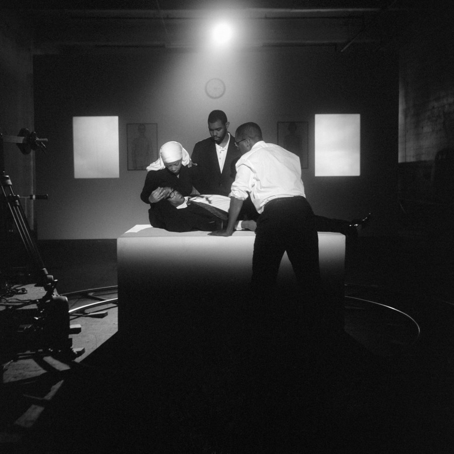 Black and white photograph showing a studio set upon which one female and two male African American figures are gathered around a supine African American man.