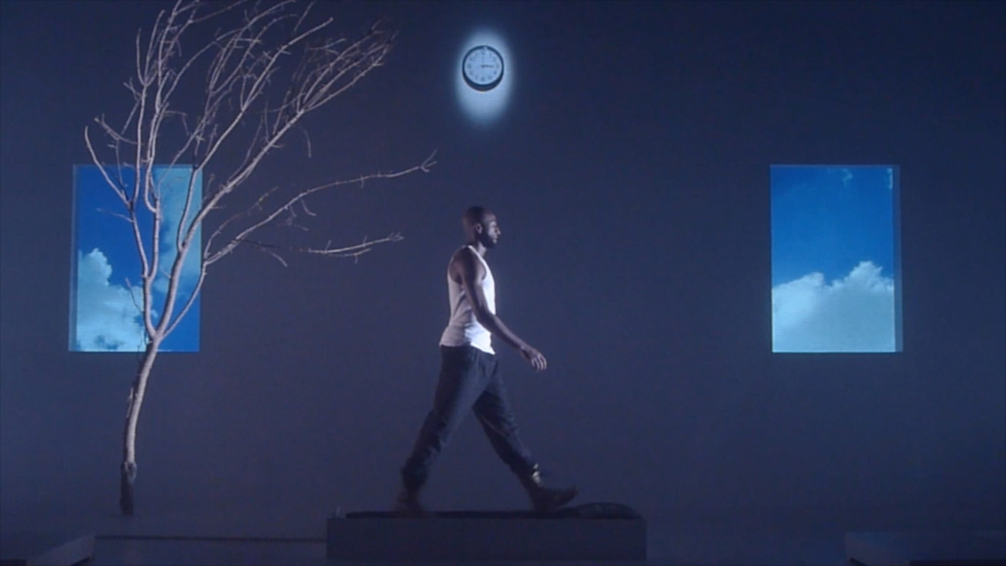 Film still from a blue-toned video depicting an African American man walking on a treadmill, flanked by a tree, two images of the sky, and a clock