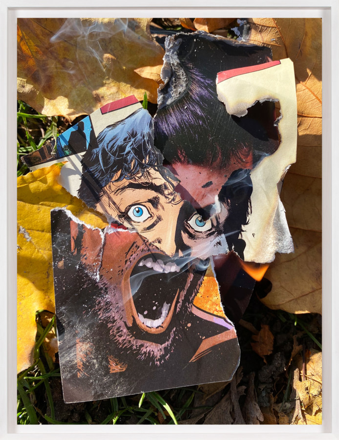 A framed color photograph of screaming faces from a comic book, torn out and lit on fire on the grass, surrounded by leaves.