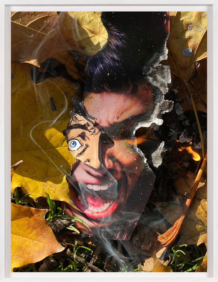 A framed color photograph of a screaming face from a comic book, torn out and lit on fire on the grass, surrounded by leaves.