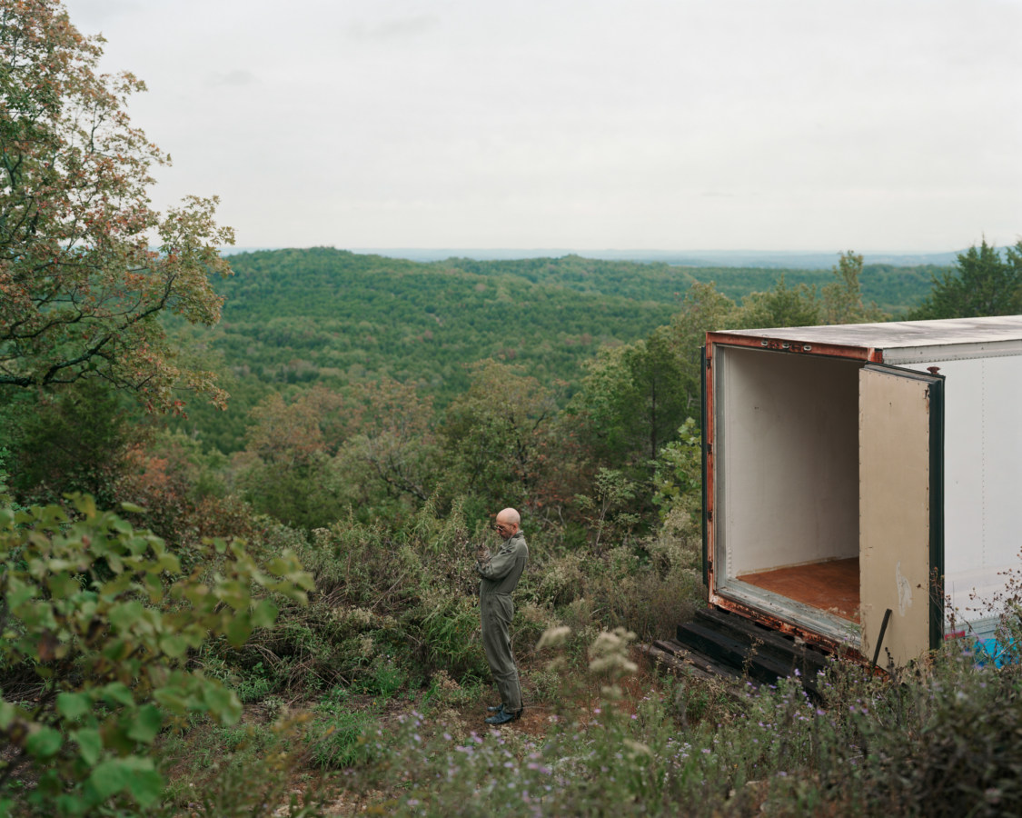 Color photograph of a man standing outside of an open box truck overlooking a green valley