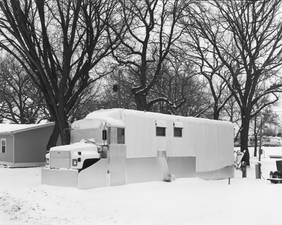 Black and white photograph of a school bus converted into a trailer home in a snowy trailer park