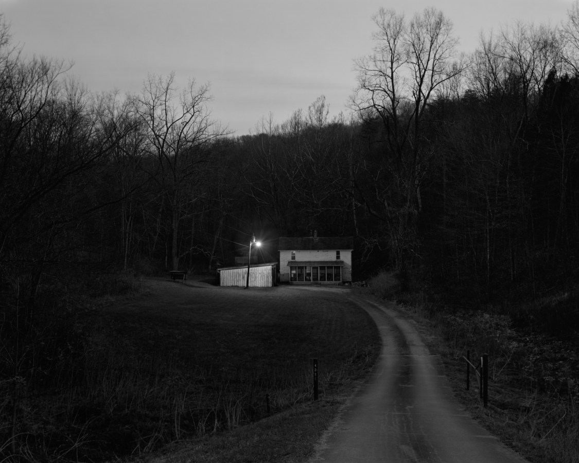 Black and white photograph of a store front and single lamppost at the end of a dirt road at dusk