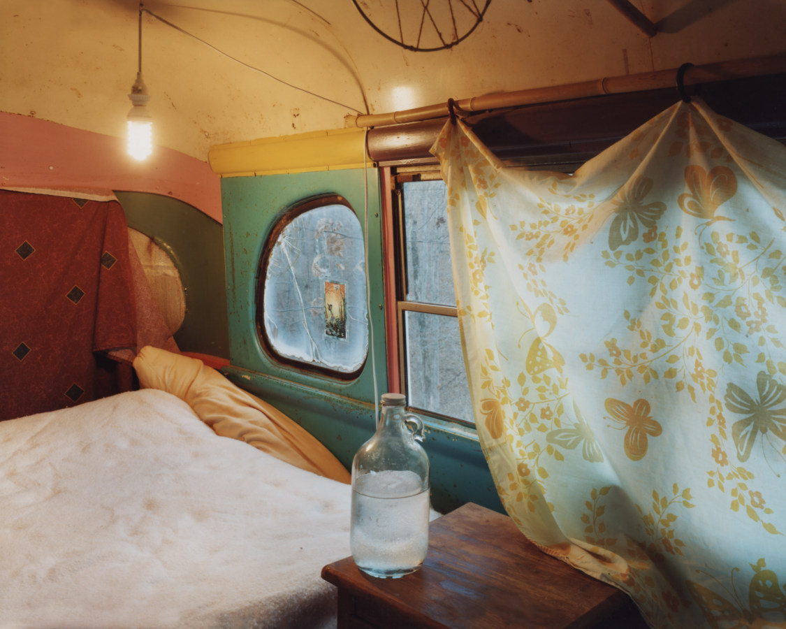 Color photograph of the interior of a mobile home with a bed and glass jug