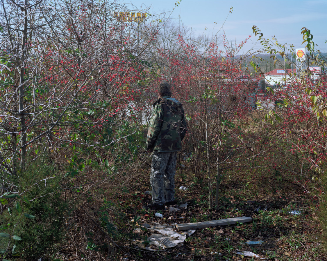 Color photograph of a man in camouflage standing amid low trees overlooking a gas station
