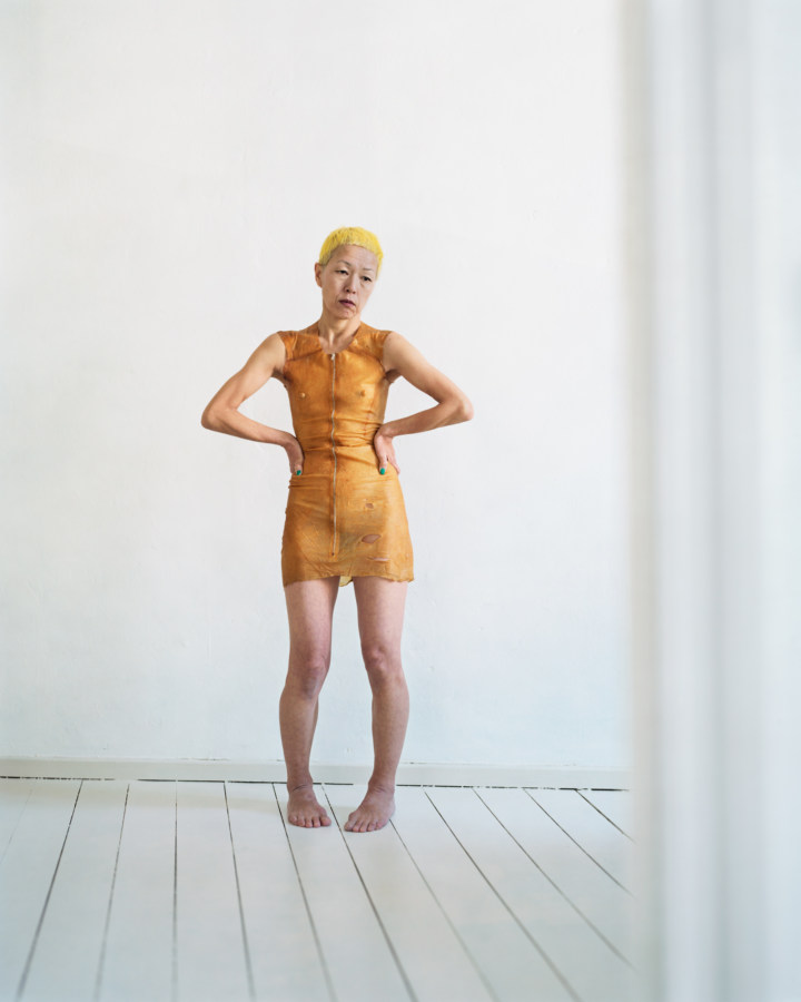 Color photograph of a woman dressed in yellow standing in an empty white room