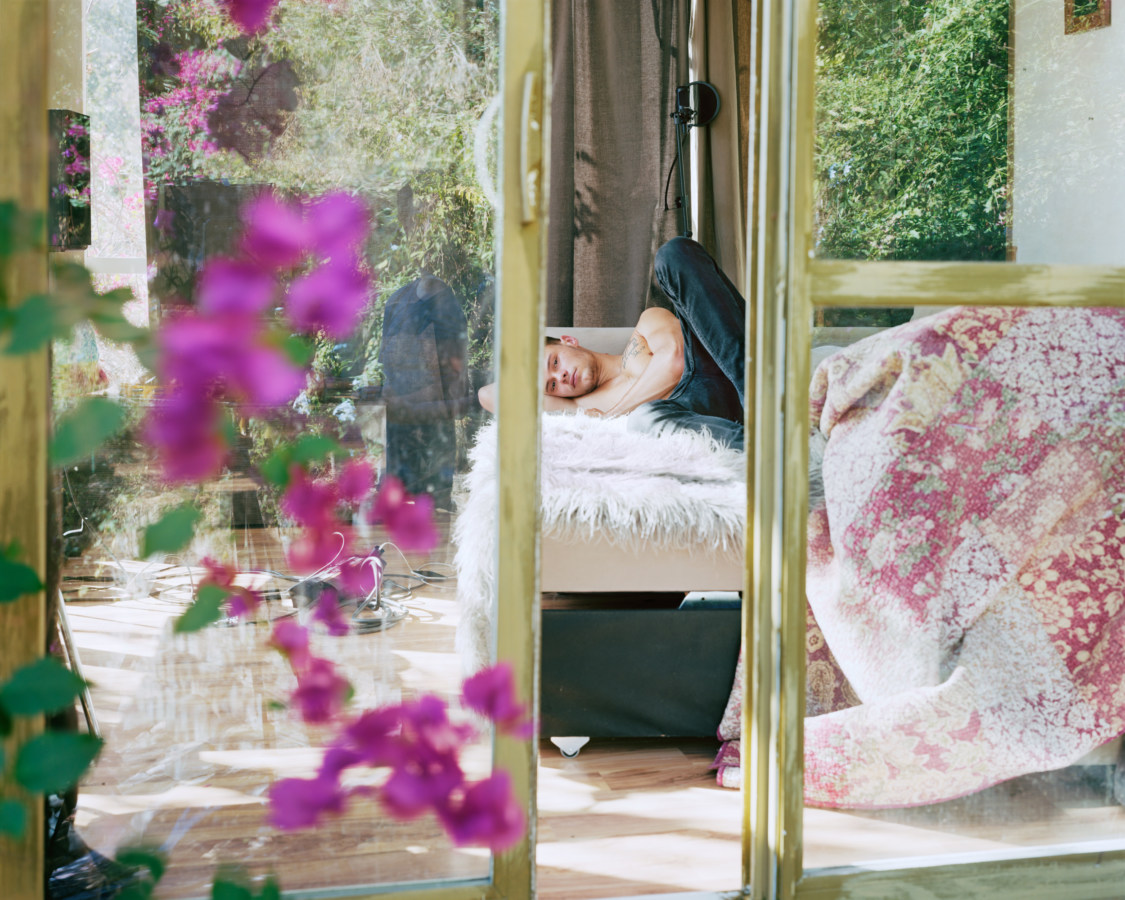 A photograph of a young man lying on a bed, as seen through a partially open sliding door. Pink flowers on the left.